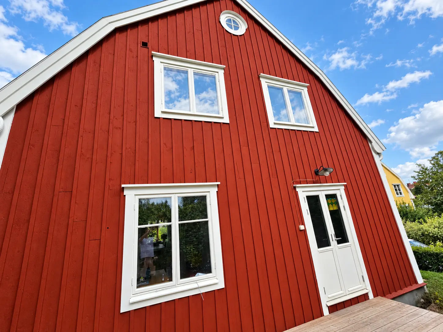 Rödmålad gavel med vita foder, dörr och trädäck framför.
